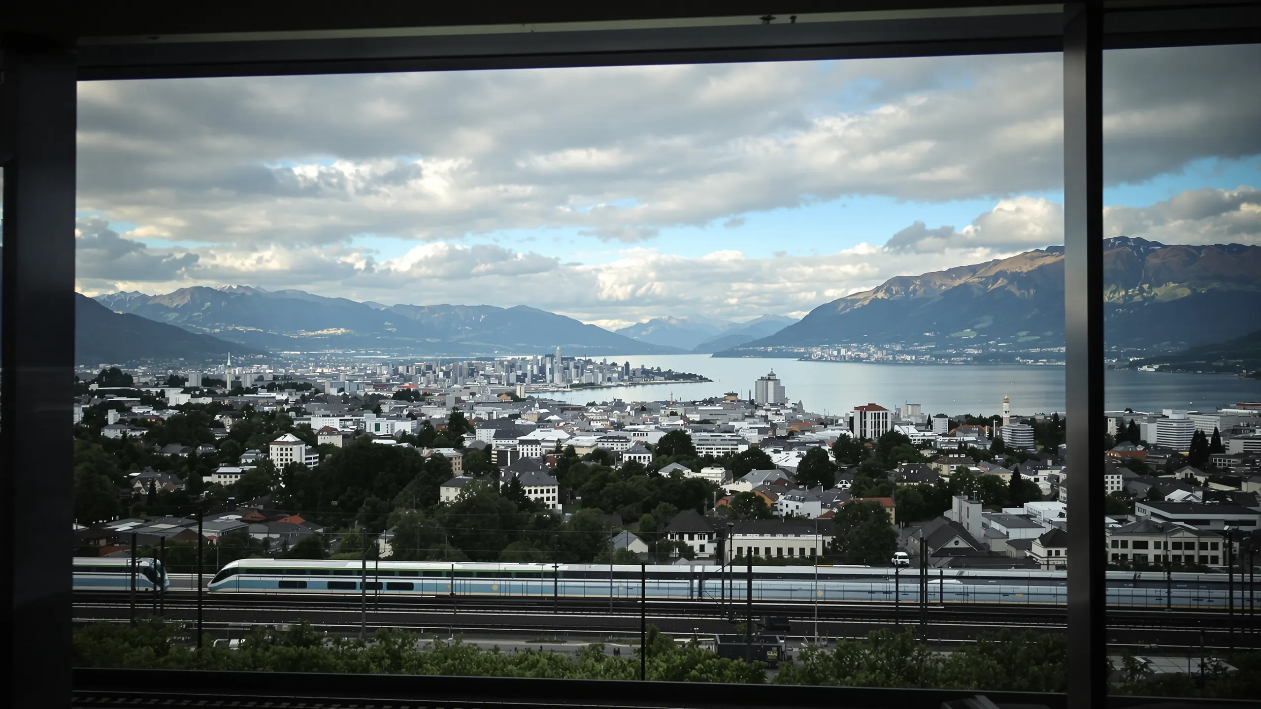 Blick durch ein Panoramafenster auf eine Schweizer Stadt mit See, Bergen und Bahnlinie